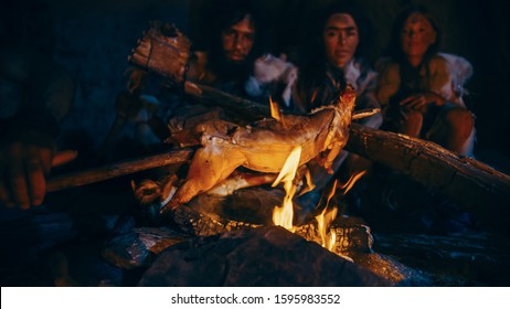 Neanderthal or Homo Sapiens Family Cooking Animal Meat over Bonfire and then Eating it. Tribe of Prehistoric Hunter-Gatherers Wearing Animal Skins Eating in a Dark Scary Cave at Night.