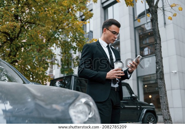 Near black car with phone in hands.\
Businessman in black suit and tie is outdoors in the\
city.