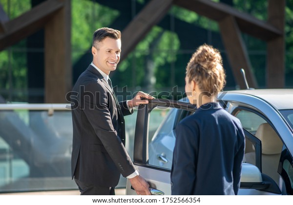 Near the car.\
Smiling man in black suit opening the door to the car and woman\
standing with her back on the\
street