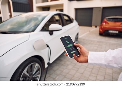 Near garage. Close up view. Woman charging her electric car and monitoring process on smartphone. - Shutterstock ID 2190972139