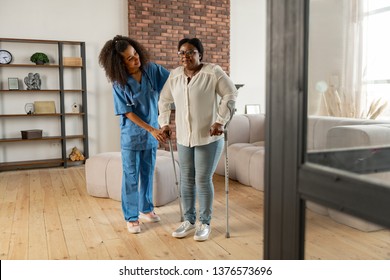 Near private nurse. Aged dark-skinned woman making first steps with crutches standing near private nurse - Powered by Shutterstock