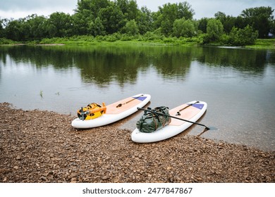 Near the rocky riverbank, two paddleboards are ready for an outdoor adventure amidst lush greenery. This calm water scene offers tranquility for nature lovers to relax and enjoy the serene setting - Powered by Shutterstock