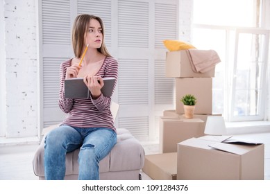 Neat and organized. Pleasant young woman sitting on the pouf in her old apartment and checking the list of items in the notebook before moving out