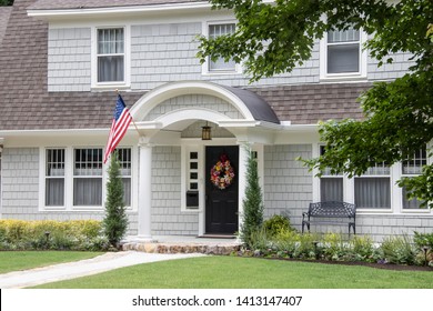 Neat And Pretty Shingled Retro House With Arched Entryway And Beautiful Landscaping With Colorful Summer Wreath On Front Door And American Flag Attached To Front Column