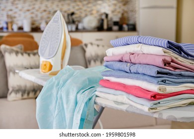 Neat Stack Of Clean Freshly Laundered Clothes And Electric Iron On Pressboard In Living Room. Pile Of T-shirts And Sweaters On Ironing Board, Home Interior Background. Copy Space, Close Up, Top View.