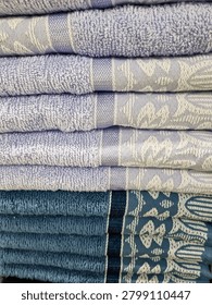 A neat stack of folded cotton bath towels in various shades of blue and soft purple. The fabric features delicate floral and geometric woven patterns, showcasing a clean, soft, and textured look suitable for bathroom or home textile concepts