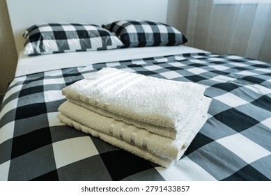 A neat stack of folded white bath towels placed at the foot of a bed dressed in a large black and white checkered duvet and matching pillows. Soft natural