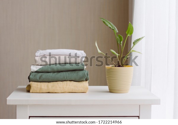 a neat stack of folded women\'s clothing in\
natural colors on a white wooden chest of drawers, a green\
houseplant near the window with white\
tulle