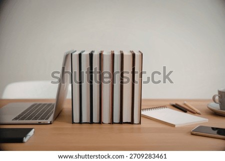 A neat stack of pastel colored notebooks stands on a wooden desk next to a laptop, notepad, and phone, suggesting a productive workspace.