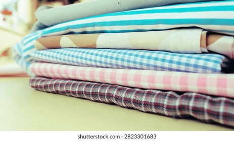 A neat stack of various colorful, patterned fabrics, including stripes, checks, and abstract shapes, is piled on a light surface. The textiles, likely cotton, are folded crisply, showcasing their different designs and hues.