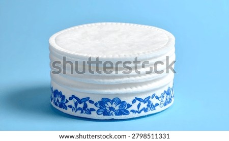 A neat stack of white cotton pads featuring a decorative blue floral border on the bottom pad, isolated on a vibrant blue background.