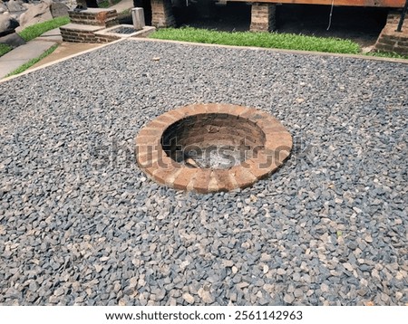 A neatly constructed circular brick firepit surrounded by a layer of gray crushed stones. The firepit sits in an open area near landscaped greenery and a rustic wooden structure.