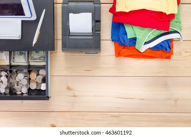 Neatly Folded Pile Of Brightly Colored Summer Clothes, An Adding Machine And Open Cash Register With American Dollar Currency On A Wooden Table In An Overhead View With Copy Space Conceptual Of Sales