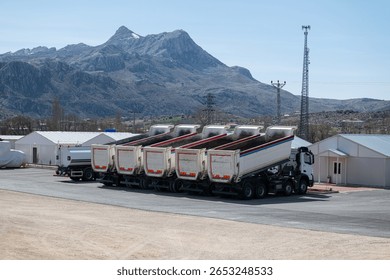 Neatly lined up in front of a facility located in a mountainous environment, raised dump trucks with tipper bodies emphasize the scale of logistics, synchronization, and transportation operations.