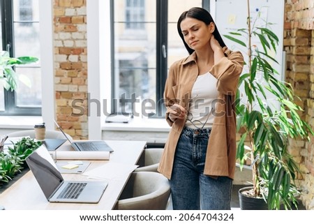 Neck ache concept. Young woman standing at office and feeling hurt having pain in her neck. Woman massaging rubbing stiff sore neck tensed muscles, fatigued from computer work and incorrect posture 