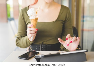 From The Neck Down View Of Young Handsome Caucasian Redhead Woman Sitting In A Bar Using Tablet And Smart Phone While Eating Ice Cream - Technology, Social Network, Multitasking Concept