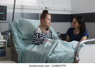 Neck Injured Hospitalized Girl With Cervical Collar Resting In Hospital Pediatric Ward. Unwell Little Kid Suffering From Car Accident Sitting In Patient Bed While Mother Comforts Her.