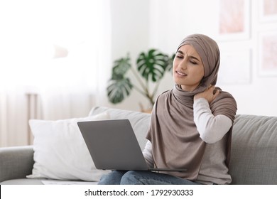 Neck Pain. Muslim Woman Massaging Inflamed Neck Tired Of Work At Laptop, Sitting On Sofa At Home, Free Space