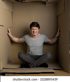 Need more space concept. Full length portrait of young depressed man is sitting in cardboard box. He is pressuring on walls while trying to extend them