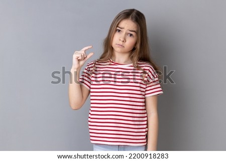 Need some more. Little girl wearing striped T-shirt doing a little bit gesture and looking with displeased imploring expression, showing minimum. Indoor studio shot isolated on gray background. Foto stock © 