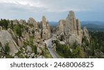 
The Needles Highway in South Dakota, with winding road passing through towering, needle-like rock formations amidst a lush forested area under a partly cloudy sky. Cars travel along the scenic route.