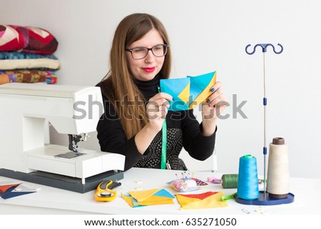 needlework and quilting in the workshop of a young tailor woman - tailor with glasses behind a desk and sewing machine considering two flaps of fabric lying next to the threads, fabrics, needles, pins
