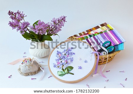 Needlework. A vase of blooming lilac and hand embroidery with satin ribbons on a white table. Sets of colored ribbons and accessories for embroidery in a wicker basket (embroidery are made by me)