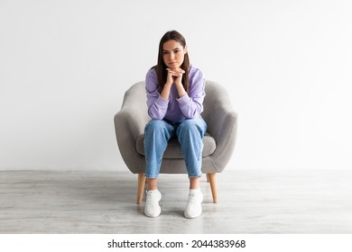 Negative emotions. Full length of young Caucasian woman sitting in armchair with dull or thoughtful face expression, feeling upset or tired, having problem or depression against white studio wall