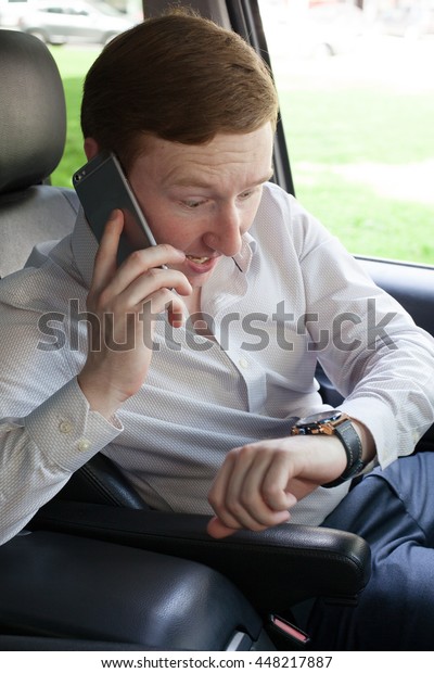Negative emotions. Late. Young man in white shirt\
sitting in the car, looks at his watch and somebody is shouting on\
the phone.