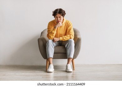 Negative emotions. Young Asian man sitting in armchair with thoughtful face expression, feeling sad or tired, having problem, suffering from depression or stress against white studio wall - Shutterstock ID 2111164409