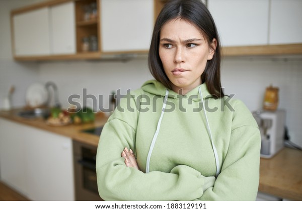 Negative human feelings and reactions. Indoor\
shot of frustrated stubborn teenage girl in hoodie biting lips,\
posing i kitchen with arms crossed, expressing disagreement, does\
not want to do\
housework