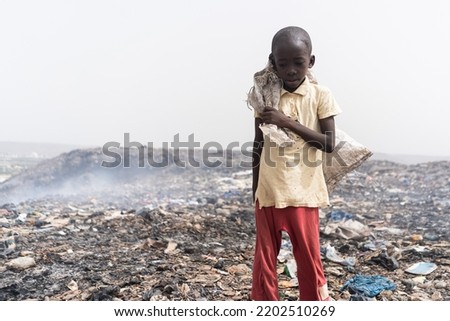 Neglected, malnourished African boy standing desolate in the midst of a smelly and smoking garbage dump; social issue of child abandon