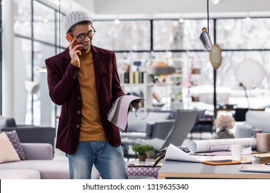 Negotiation. Good-appealing brown-haired smiling procurement specialist in his 30s wearing fancy blazer, grey hat and glasses negotiating on phone with fabrics supplier while holding samples in hands