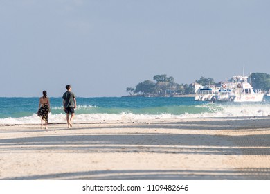 Negril, Jamaica - March 24 2018: Young Millennial Caucasian Woman And Man Couple Taking Morning Walk On Seven Mile Beach In Negril. Yachts/Boats Docked On Ocean. High Tide Waves Crashing On Shore.