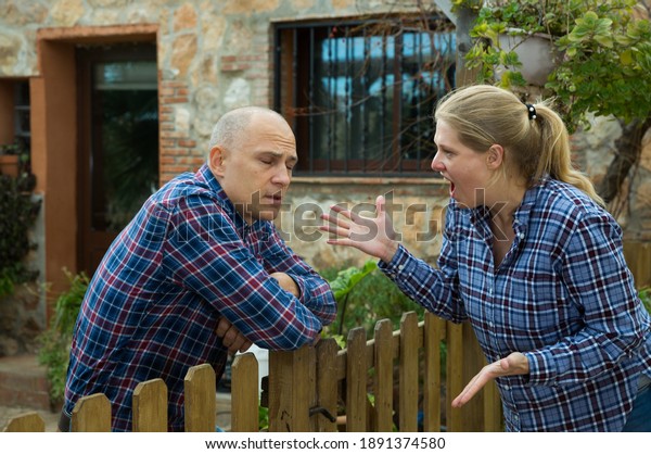 Neighbor quarrel. Irritated woman\
and man emotionally talking near wooden wicket of rural\
house