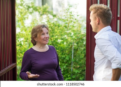 Neighbors Discuss The News, Standing At The Fence. An Elderly Woman Talking With A Young Man. They Are Satisfied With This Meeting