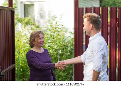 Neighbors Discuss The News, Standing At The Fence. An Elderly Woman Talking With A Young Man. They Are Satisfied With This Meeting