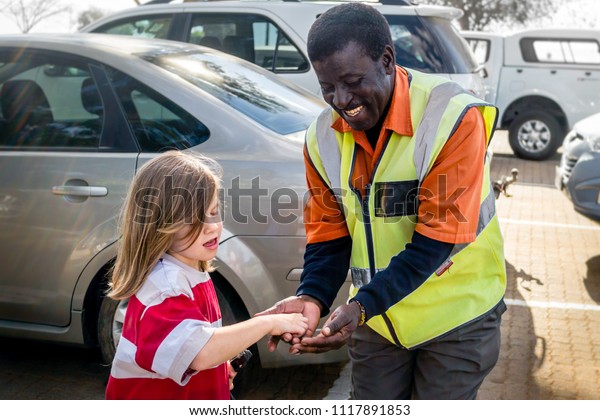 NELSPRUIT, SOUTH AFRICA – JULY 30, 2013:
A young white girl tips a friendly black security car park
attendant.  Car guards are prevalent in South Africa and have
controversial
self-employment.
