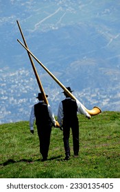 Nendaz, Conthey District, 
Valais, Wallis, Switzerland, Europe - two men in traditional black vests, trousers and hats walking with alphorn on their arms, Swiss Alps, Nendaz village seen in valley - Shutterstock ID 2330135405