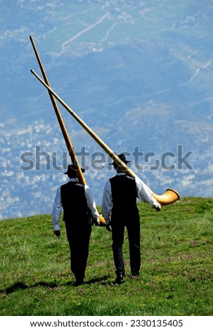 Nendaz, Conthey District, 
Valais, Wallis, Switzerland, Europe - two men in traditional black vests, trousers and hats walking with alphorn on their arms, Swiss Alps, Nendaz village seen in valley