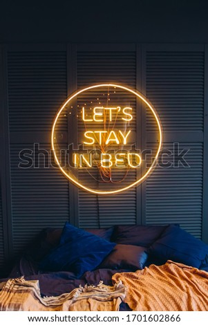 Neon inscription in the bedroom on the wall. Light sign - let's stay in bed. Quarantine during a coronavirus pandemic. Compliance with self-isolation.