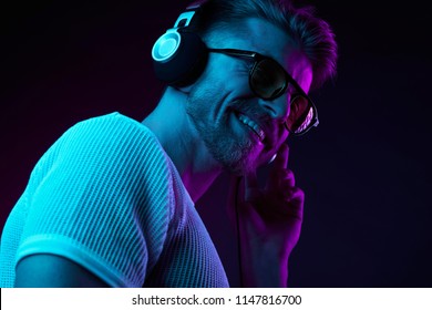 Neonový lehký portrét vousatého úsměvného muže ve sluchátkách, sluneční brýle, bílé tričko. Poslech hudby