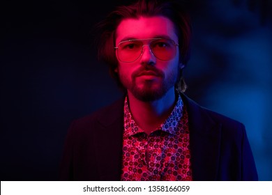 Neon Light Portrait Of Handsome Male Model With Dark Medium Length Hair, Mustaches And Beard In Sunglasses, Weared In Dark Jacket And Floral Shirt.