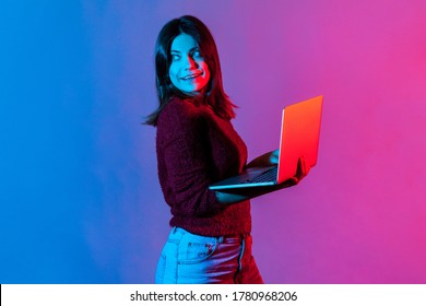 Neon Light Portrait Of Successful Joyful Office Worker Girl Looking Away With Happy Pensive Face, Smiling Holding Laptop, Typing On Keyboard, Doing Freelance Job. Colorful Neon Light, Studio Shot