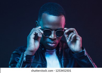 Neon Light Studio Portrait Of African American Male Model Wearing Trendy Sunglasses And Leather Jacket