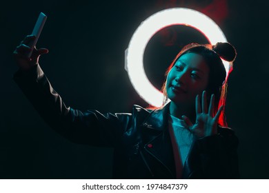 Neon Night Selfie. Cyberpunk Life. Future Communication. Blue Red Color Light Smiling Asian Girl In Black Leather Jacket With Round Halo Waving Hand On Phone Camera On Dark.