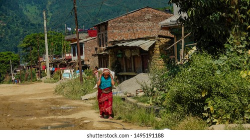 NEPAL COUNTRYSIDE, OCTOBER 2018: Older Asian Woman Carries A Heavy Bag On Her Back Through The Poverty Stricken Village In Nepal. Senior Woman Walks Past Young Girl While Carrying A Heavy Bag Of Grain