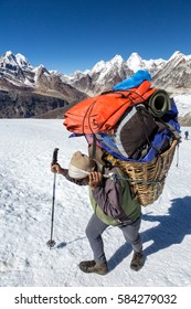 Nepalese Sherpa Porter Walking On Glacier With Trekking Pole Carrying Basket With Lots Of Mountain Expedition Luggage Using Traditional Head Strap In Himalaya