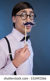 Nerd Making A Face. Young Nerd Man Holding A Stick With Fake Moustache At His Face While Isolated On Blue