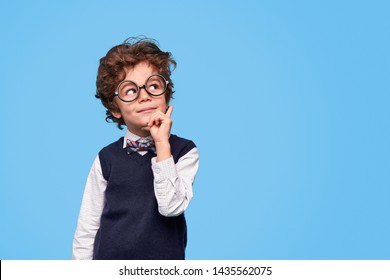Nerdy boy in round eyeglasses and school uniform rubbing chin and looking away while thinking near empty space against blue background - Shutterstock ID 1435562075